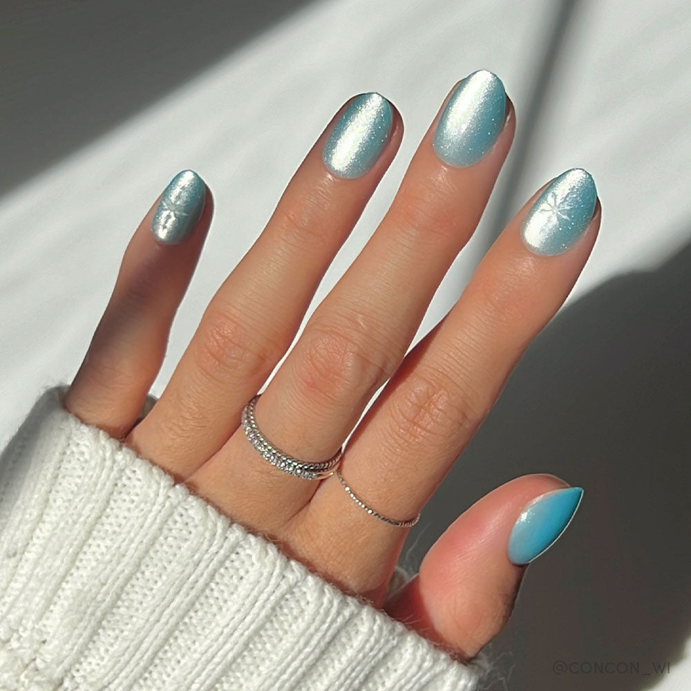 A hand with glamnetic’s Subzero nails in light blue and silver chrome, detailed with a snowflake accent, is styled with silver rings and a white knit sweater. Soft lighting casts gentle shadows in the background. - Product image