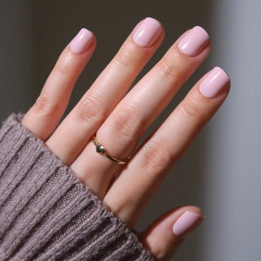 A hand with super short nails painted in glamnetic’s Sugar BB classic pink, wearing a simple gold heart ring and a mauve ribbed sweater sleeve partially covering the wrist. - Product image
