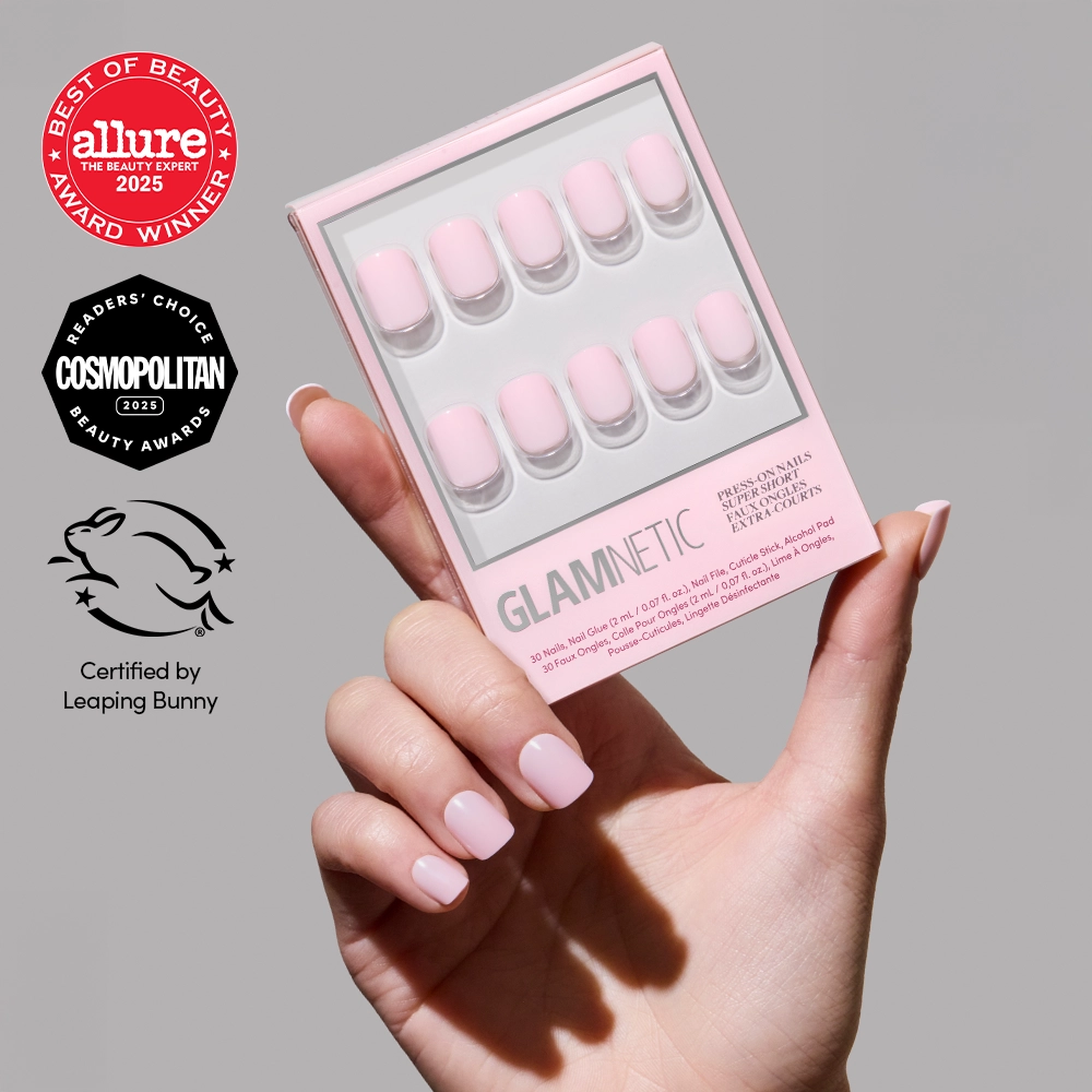 A hand with a classic pink mani of glossy Sugar BB press-on nails by glamnetic holds a box of similar super short nail press-ons. The image displays Allure, Cosmopolitan, and Leaping Bunny certification logos. - Product image
