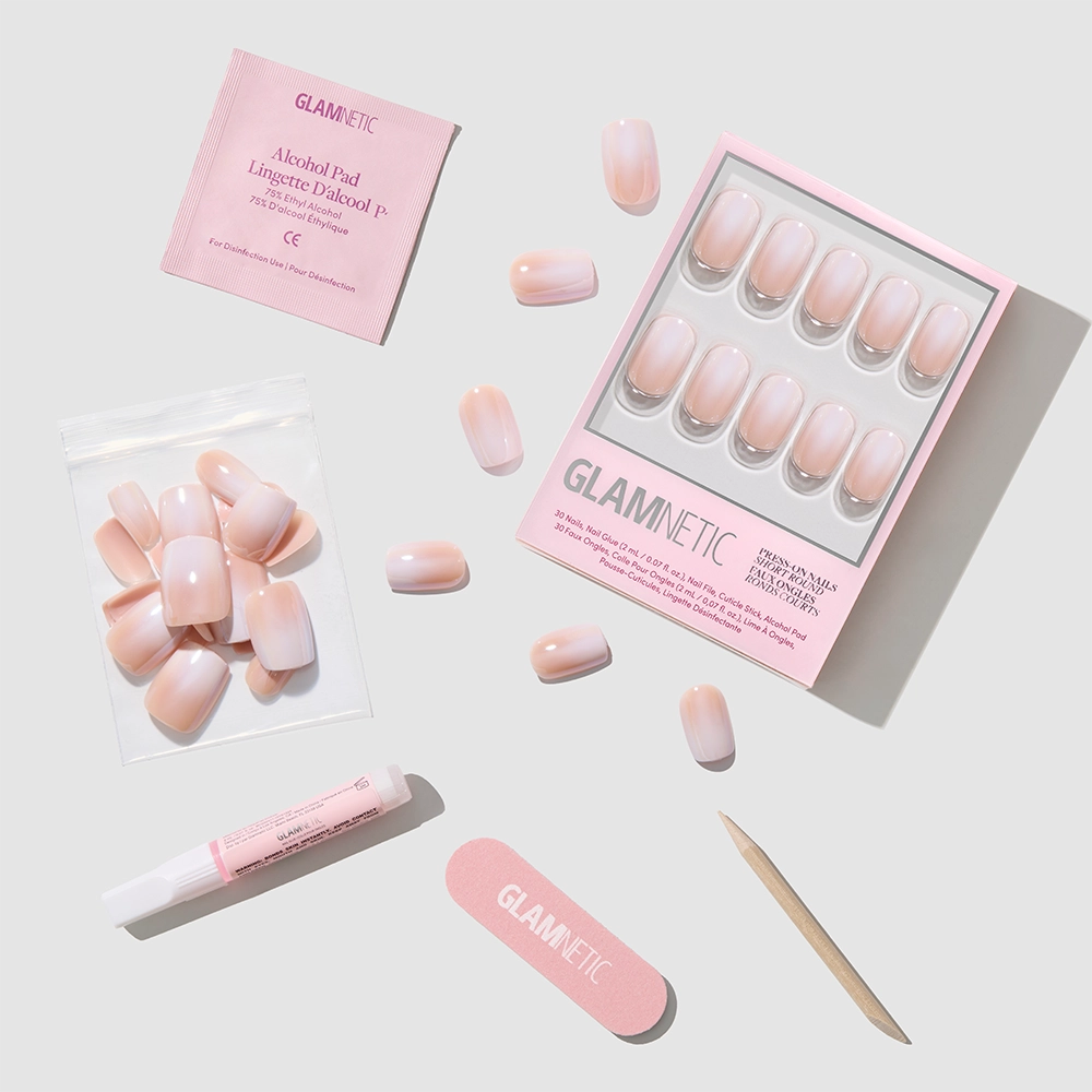 Sugar Veil Press-On Nails by glamnetic, featuring ombré French tips, comes with nail glue, cuticle stick, nail file, alcohol pad, and extra nails—arranged on a light gray background beside branded packaging. - Product image