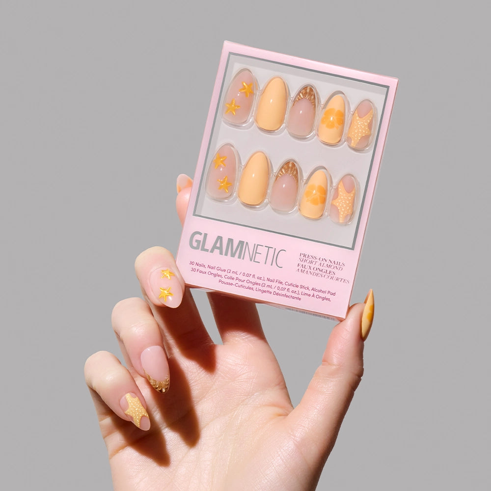 A hand with patterned yellow press-on nails holds the glamnetic Sunny Shores Press-On Nail Set, featuring yellow and nude designs with stars, abstract shapes, and seashell details for a mermaid-inspired look against a plain grey background. - Product image