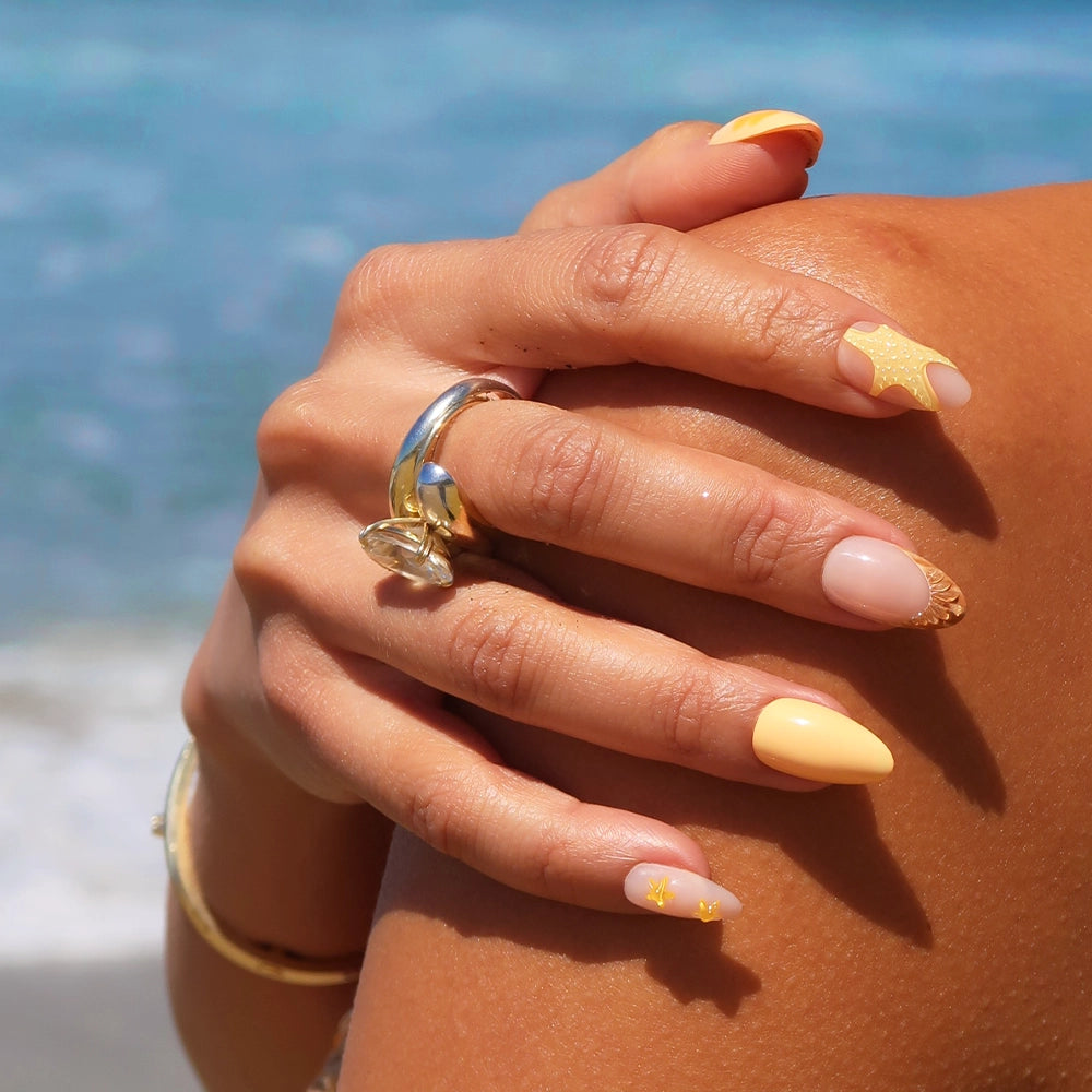 A close-up of hands wearing glamnetics Sunny Shores almond-shaped nails in yellow and nude, with gold star and seashell accents, resting together. Gold rings and bracelets adorn the hands, with a blurred ocean background. - Product image