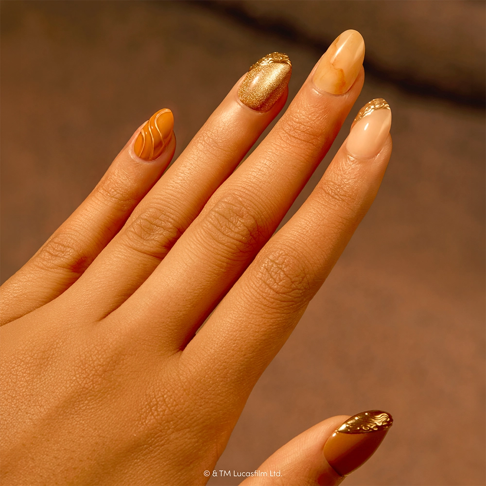 A hand with long, almond-shaped nails painted in gold and nude shades, featuring intricate gold designs and marbled patterns on some nails—the Suns of Tatooine nail set by glamnetic draws inspiration from the desert planets of STAR WARS. This look stands out beautifully against a blurred brown background, blending elegance with a touch of galactic flair. - Product image