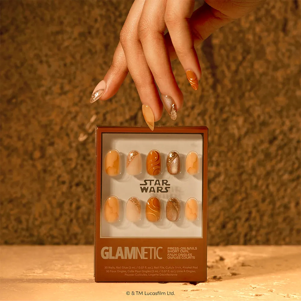 A hand with long painted nails holds a glamnetic Suns of Tatooine nail set box, featuring brown, gold, and tan Star Wars-inspired designs, set against a golden-brown textured background. - Product image