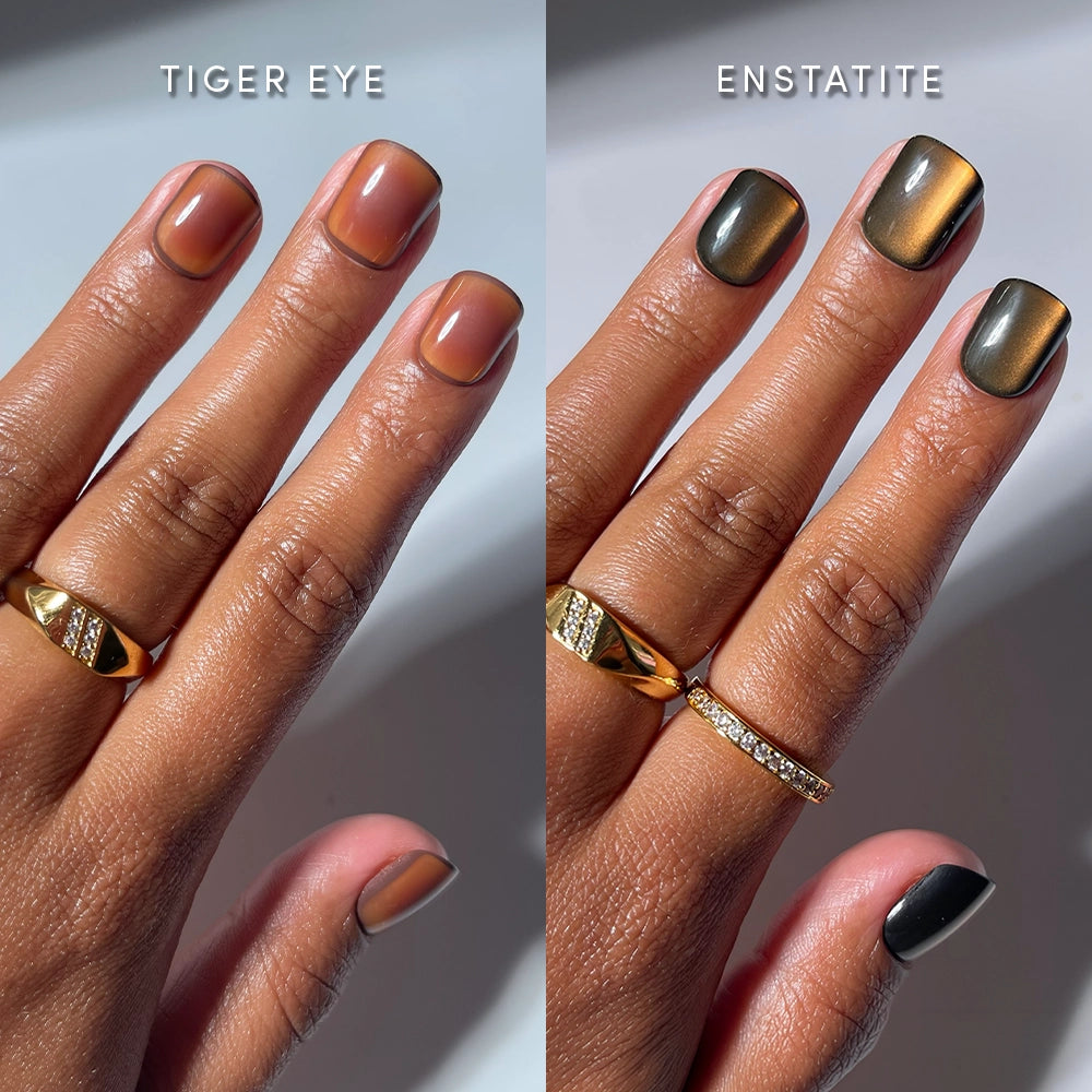 Two hands with manicured nails: the left showcases the rich brownish "Tiger Eye" from glamnetic’s Super Short Frequencies collection, while the right features a shimmery dark "Enstatite" shade. Both hands are adorned with gold rings, with each nail color labeled above its respective hand. Inspired by gemstones, these glamnetic press-ons deliver salon-quality wear for a flawlessly polished look.