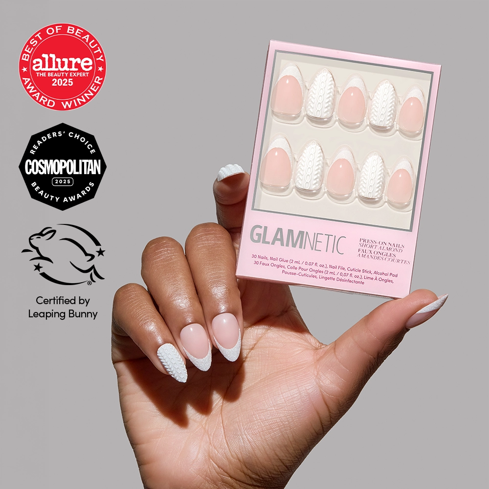 A hand holds a box of glamnetic Sweater Weather press-on nails, featuring pink and white glitter French tips, with Allure, Cosmopolitan, and Leaping Bunny certification badges visible on the gray background.