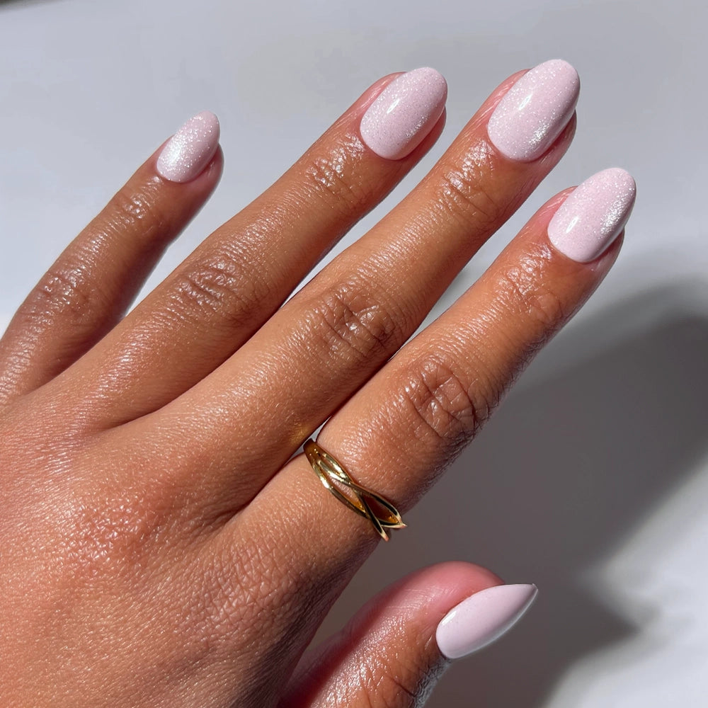 A hand with neatly manicured, short oval nails painted in glamnetic’s Sweet Talk, a shimmery pale pink, wears a simple gold twisted ring on the middle finger. Set against a plain white background, perfect for highlighting these elegant nails.