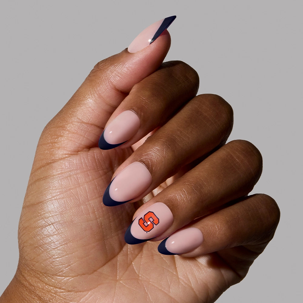 A hand with almond-shaped nails wearing the glamnetic Syracuse Orange Press-On Nail Set, featuring a nude base, navy blue French tips, and an orange S logo on the ring finger—set against a plain light gray background. Perfect for game day!. - Product image