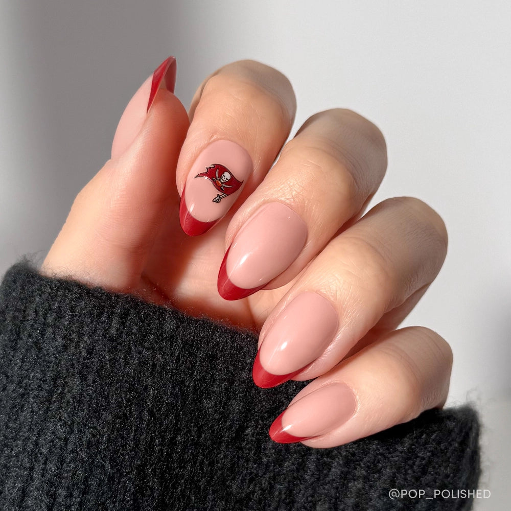 A hand wears glamnetic's Tampa Bay Buccaneers Press-On Nail Set—glossy almond French tips with a nude base, bold red edges, and a pirate flag accent—styled with a black knit sweater.