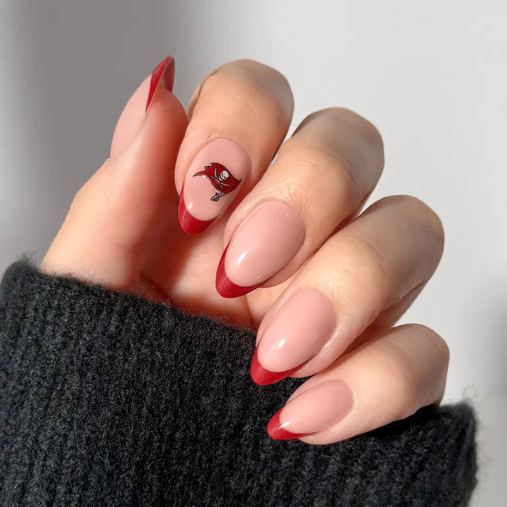A hand showcases almond-shaped NFL Press-On Nails by glamnetic, styled with nude polish and dark red French tips; one nail displays a small pirate flag accent. The person is wearing a dark, fuzzy sweater. - Product image