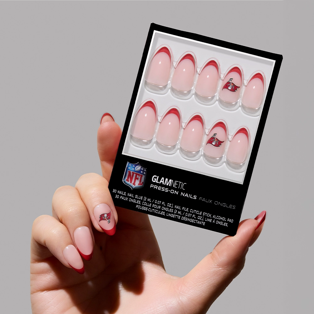 A glossy manicured hand holds a glamnetic Tampa Bay Buccaneers Press-On Nail Set, featuring almond French tips in red with the team's logo on select nails.