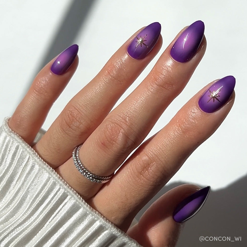 A hand with short almond-shaped nails painted in glamnetics Tarot mystical purple, featuring gold star decals on two nails. A silver ring and white ribbed sleeve partially cover the hand. - Product image