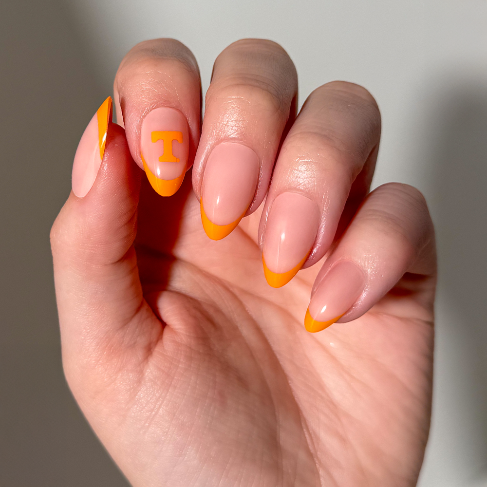 A hand with almond-shaped Collegiate Press-On Nails by glamnetic, featuring a nude base and vibrant orange French tips. One nail displays an orange “T” design near the cuticle. - Product image
