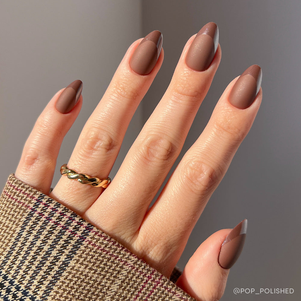 A hand with glamnetic Terra almond-shaped nails in earthy brown, featuring a matte finish and glossy brown half-moon French tips, is accessorized with a gold twisted ring and paired with a plaid sleeve. - Product image