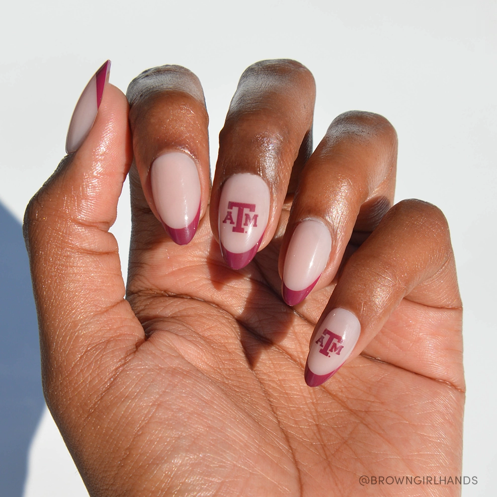 A hand displays the glamnetic Texas A&M Aggies Press-On Nail Set: almond-shaped nude nails with maroon French tips, plus two accent nails featuring the Texas A&M University logo in maroon on a white base.