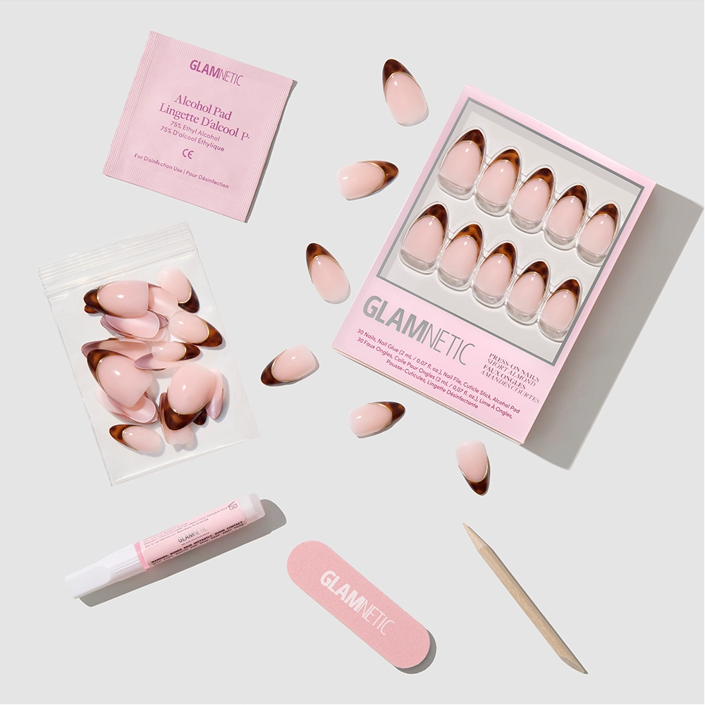 The Original by glamnetic: almond-shaped press-on nails with pink and gold French tips, shown with a nail file, cuticle stick, glue tube, alcohol pad, and extra nails on a light-colored surface. - Product image