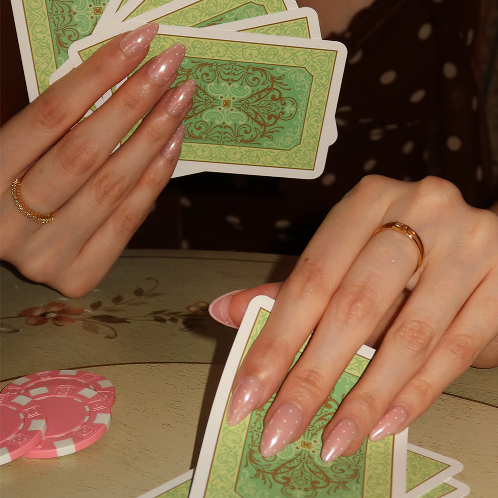 A person wearing glamnetic’s Barely Spotted nails—long, pale pink with a glazed finish—holds green-backed playing cards above a pink poker chip while dressed in a brown polka-dot top. - Product image