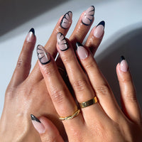 A close-up showcases glamnetic's Butterfly Effect nails—short almond shape with black French tips and abstract black lines. Gold rings adorn three fingers while the out-of-focus background highlights the elegant velvet nude design.
