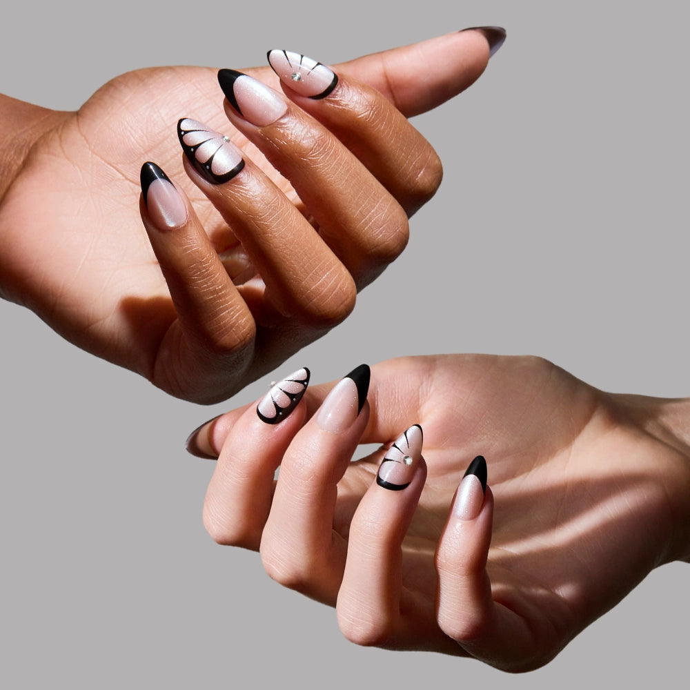 Two hands display glamnetic’s Butterfly Effect press-ons: short almond nails with a velvet nude base, black French tips, butterfly designs, and rhinestone accents against a gray background. - Product image