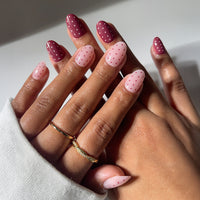 A close-up of hands wearing glamnetic’s Cute As Cherry Pie vegan, cruelty-free nails—almond-shaped in alternating pink and burgundy with red and pink polka dots. Two gold rings and a white long-sleeve shirt are also shown.