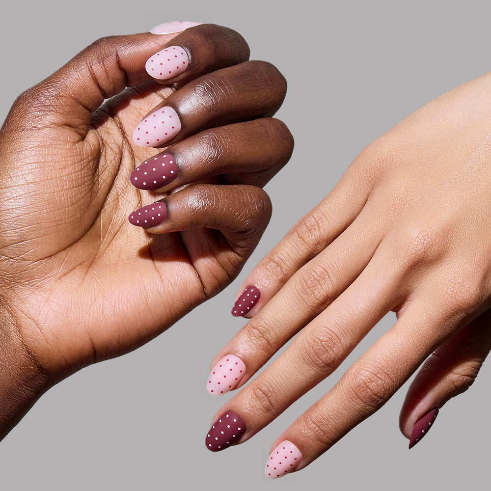 Two hands with manicured nails wear glamnetic’s Cute As Cherry Pie short oval press-ons, painted in alternating light pink and mauve with tiny polka dots, set against a plain gray background. - Product image