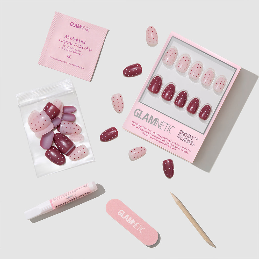 A flat lay of glamnetic Cute As Cherry Pie short oval press-on nails kit, with red and pink polka dot nails, nail glue tube, pink nail file, cuticle stick, alcohol pad, and extra nails in a bag on a light background. - Product image