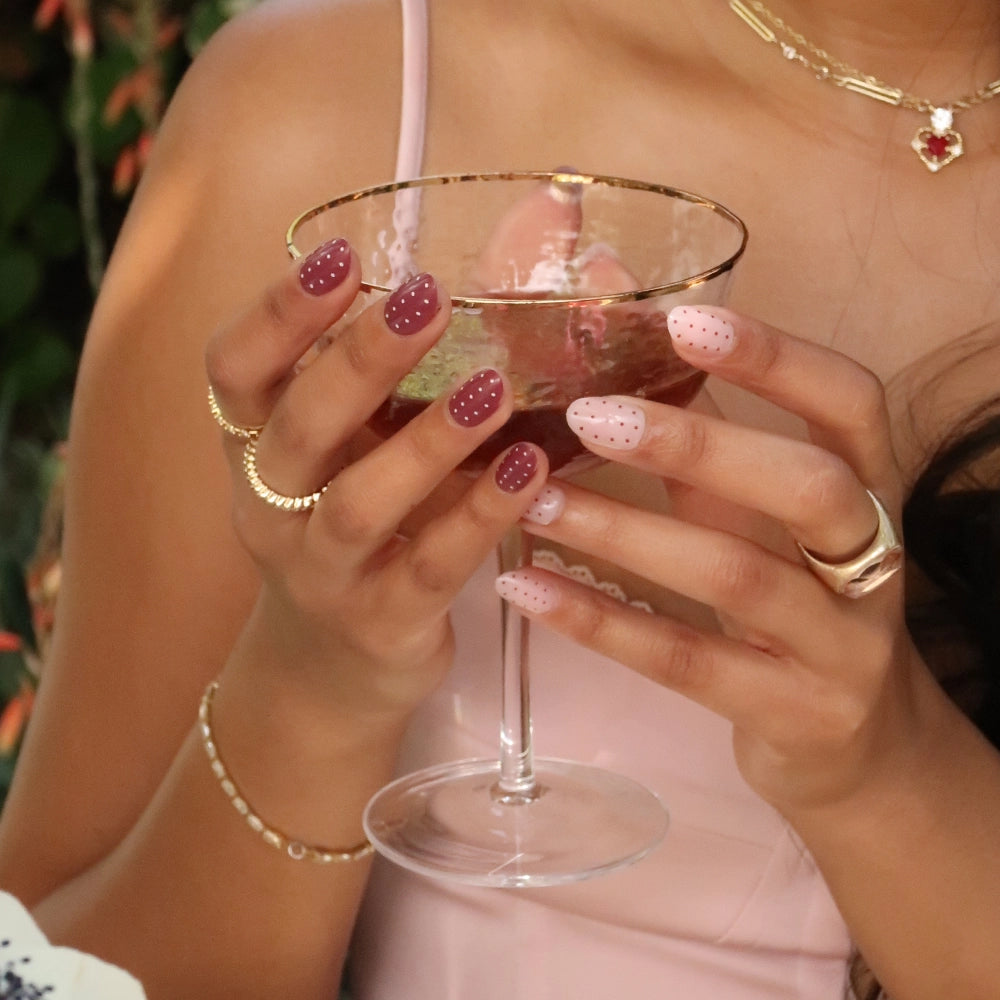 A woman in a pink dress with gold jewelry holds a gold-rimmed glass. She wears glamnetic's Cute As Cherry Pie vegan, cruelty-free nails in mauve and pale pink with polka dots, and a necklace with a red heart pendant is visible. - Product image