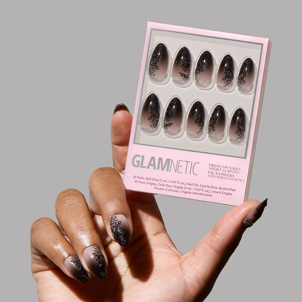 A hand with long black almond nails, lace accents, and floral designs holds a pink box of glamnetics ‘Til Death Nail Set, featuring matching black floral patterns on a nude background.