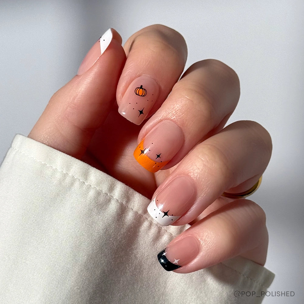 The glamnetic Tiny Pumpkin set showcases short, square Halloween nails with a pumpkin design, black and white tips, ghost nail art, tiny black stars, and an orange accent—chic against a beige sleeve. - Product image