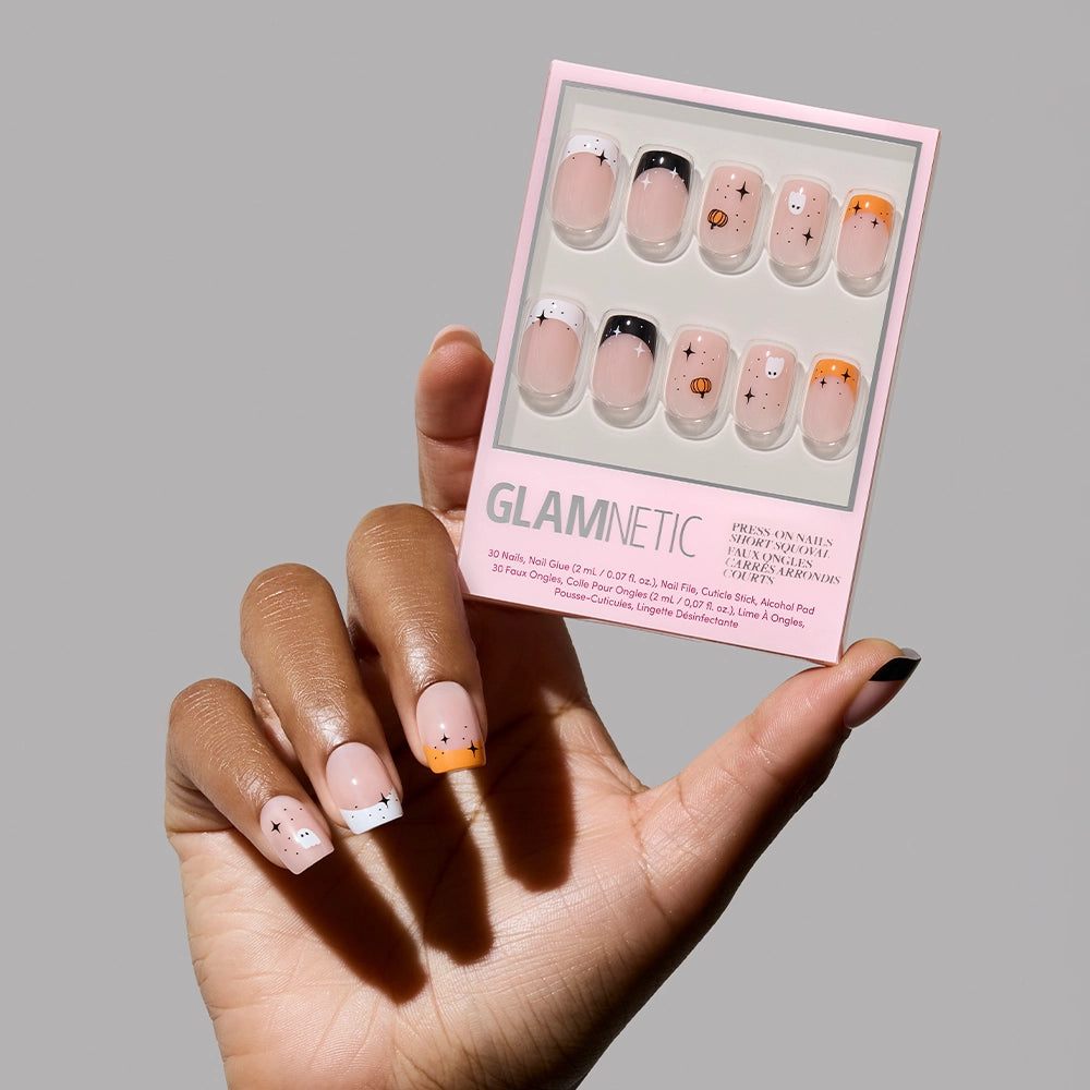 A hand with patterned press-on nails holds a box of glamnetic Tiny Pumpkin press-on nails, showcasing Halloween designs like small stars, moons, faces, and ghosts on neutral and orange backgrounds.
