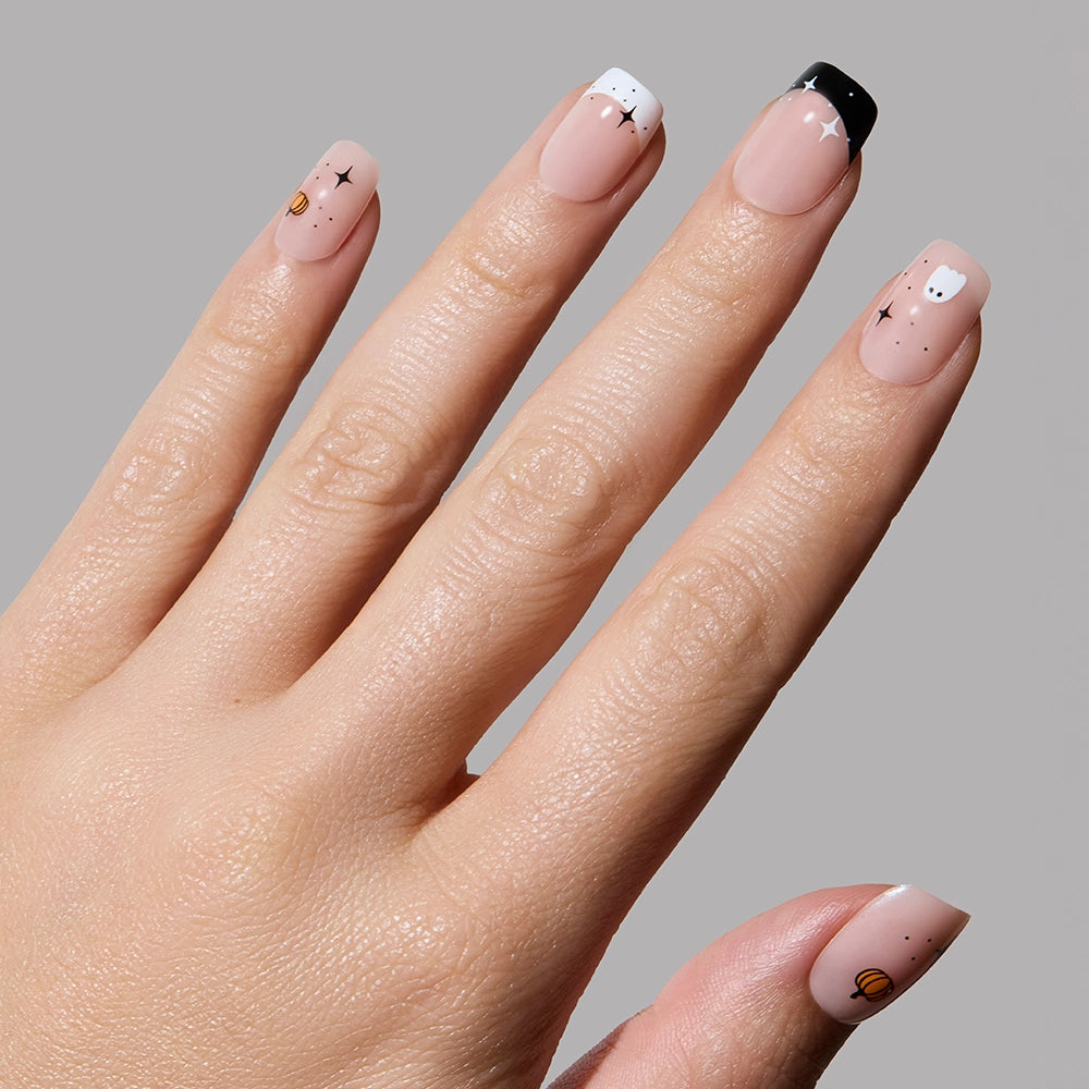 A hand with glamnetic’s Tiny Pumpkin features light nude Halloween nails with minimalist ghost, star, and pumpkin designs in white, black, and orange on a gray background. - Product image