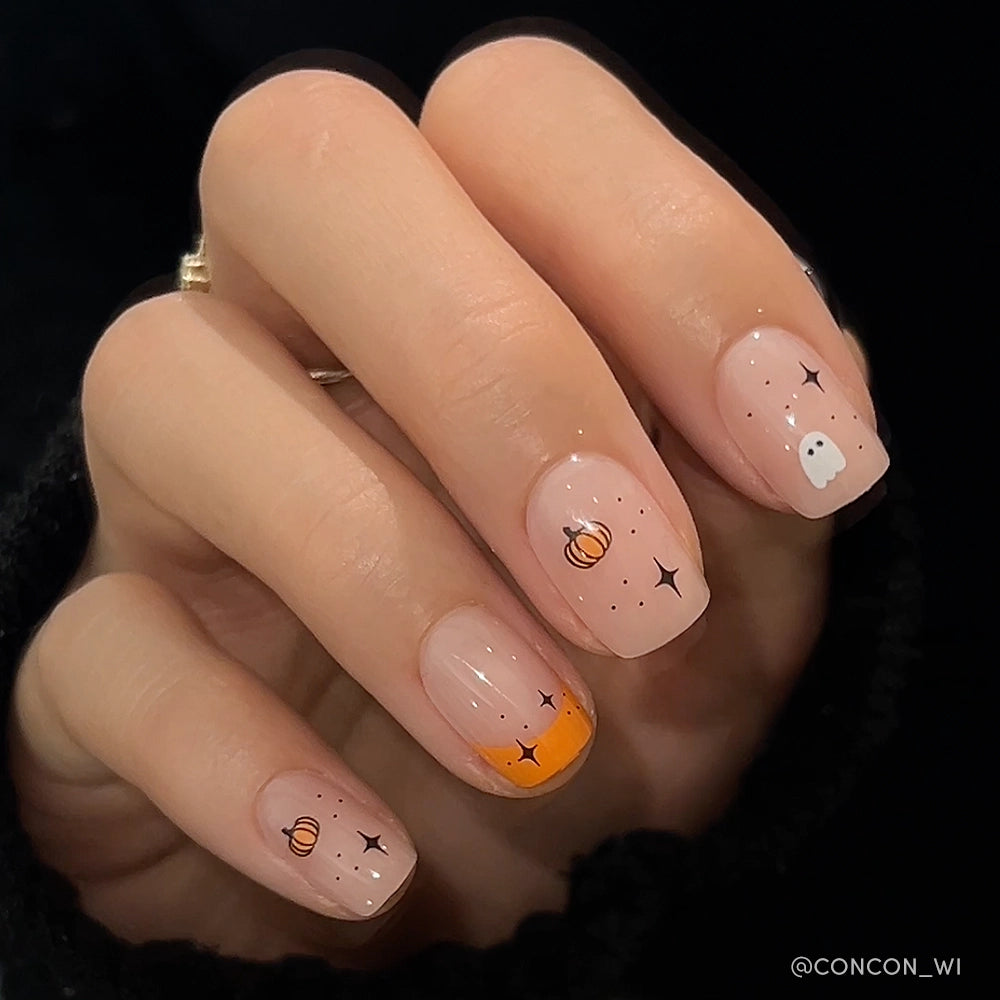 A hand with glamnetic Tiny Pumpkin Halloween nails—tiny pumpkins, stars, and ghosts on a nude base, plus one orange-tipped nail. The person wears a black sweater. - Product image