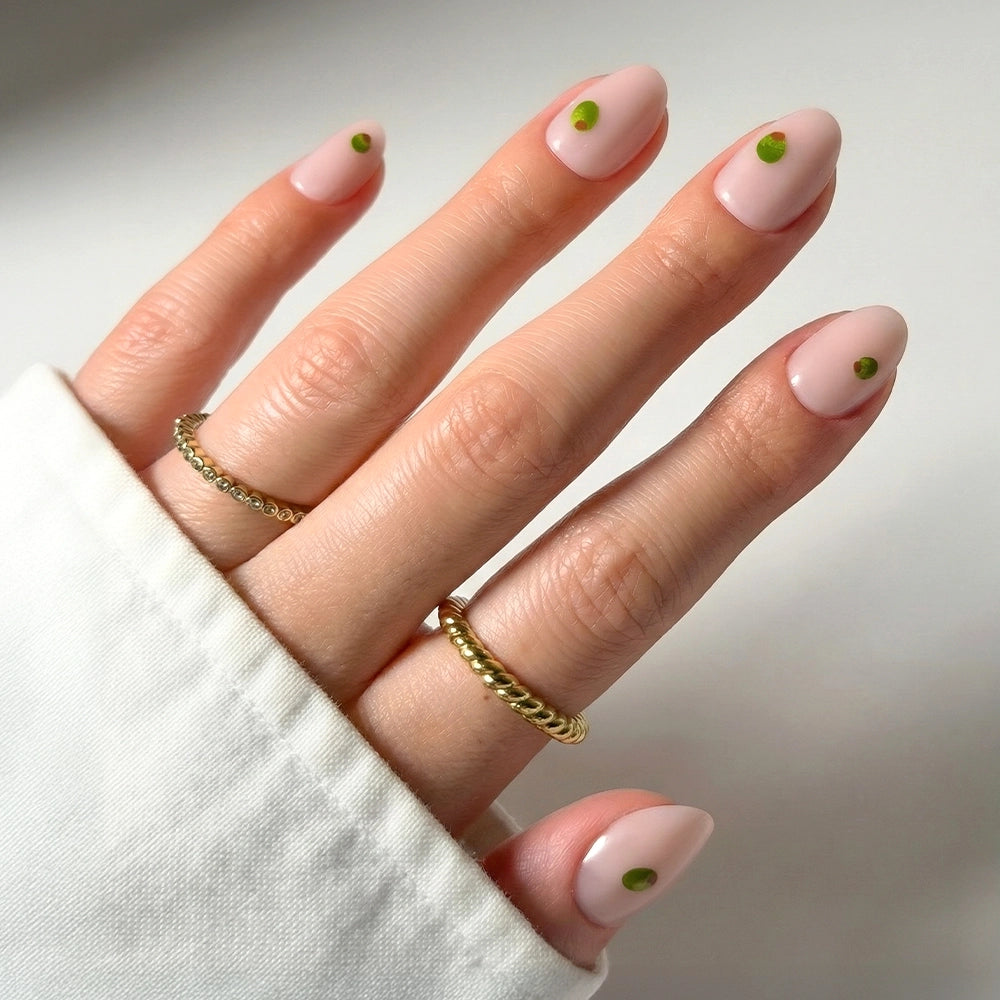 A hand with glamnetic Tipsy Olive Short Oval Nails painted light pink, each showing a green dot near the cuticle. Two gold rings are worn, and a white sleeve partially covers the hand.