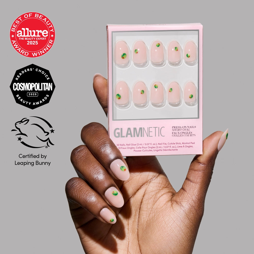 A hand with Short Oval Nails in the "Tipsy Olive" design by glamnetic, featuring green dot accents, holds a matching box of glamnetic Press-On Nails. Allure, Cosmopolitan, and Leaping Bunny certification logos are visible.