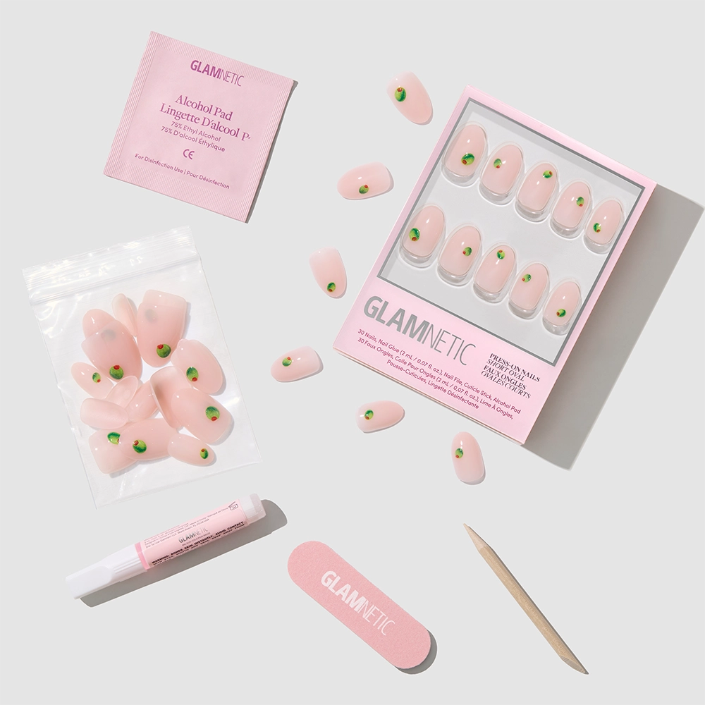 The glamnetic Tipsy Olive set features extra short oval pink press-on nails with green olive designs, a pink nail file, wooden cuticle stick, nail glue, and an alcohol pad—all neatly arranged on a light gray surface. - Product image