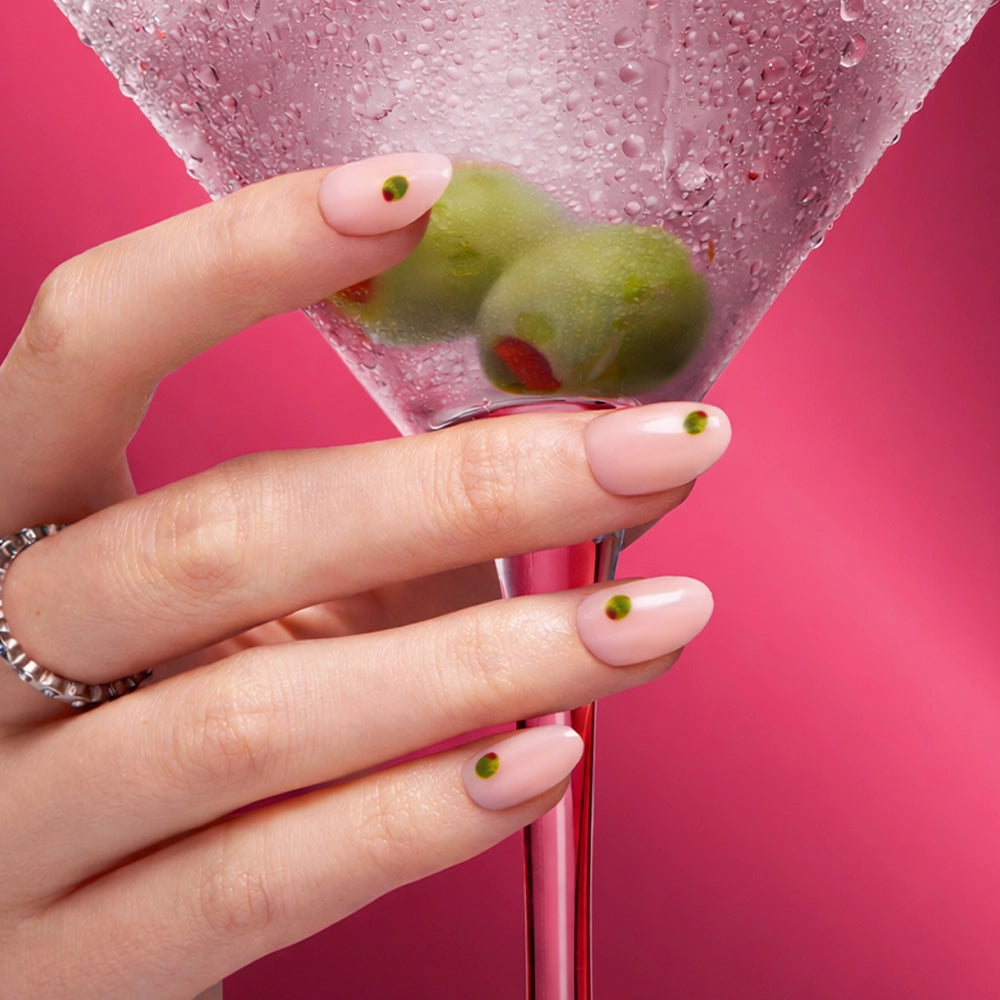 A hand with the glamnetic Tipsy Olive short oval nails in pale pink and olive accents holds a martini glass with green olives. A silver ring and bright pink background add flair, as water droplets glisten on the glass. - Product image