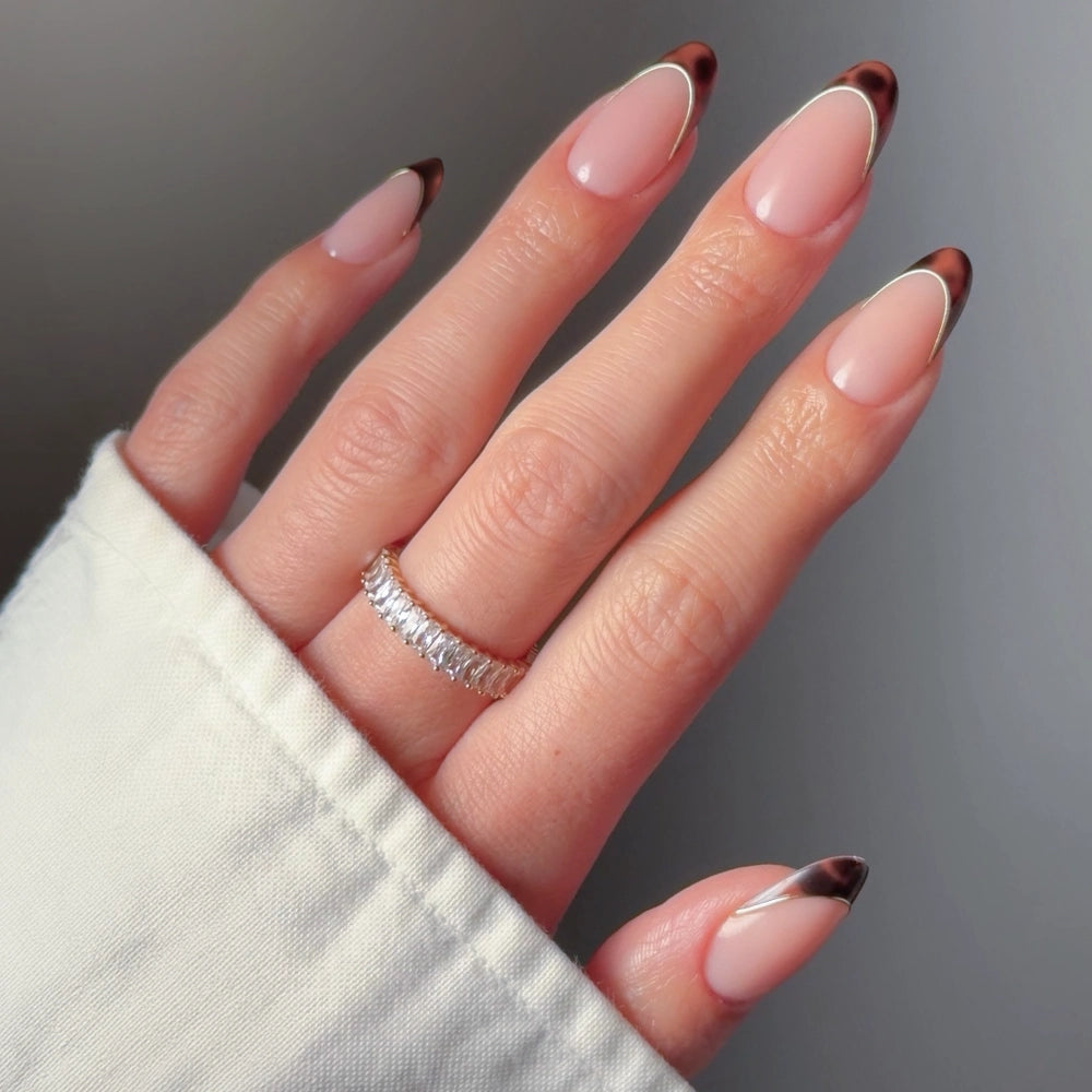 A hand showcases glamnetic's "Too Much" oval press-on nails with a nude base, brown tips outlined in white, paired with a diamond-encrusted ring and a white long-sleeve top. - Product image