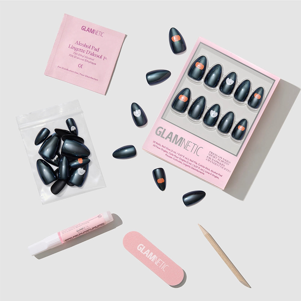 Flat lay of the glamnetic Tricks & Treats Press-On Nail Set, featuring black cat-themed short almond nails with a glazed finish, shown alongside nail glue, alcohol pad, nail file, wooden cuticle stick, and extra nails on a light gray background. - Product image