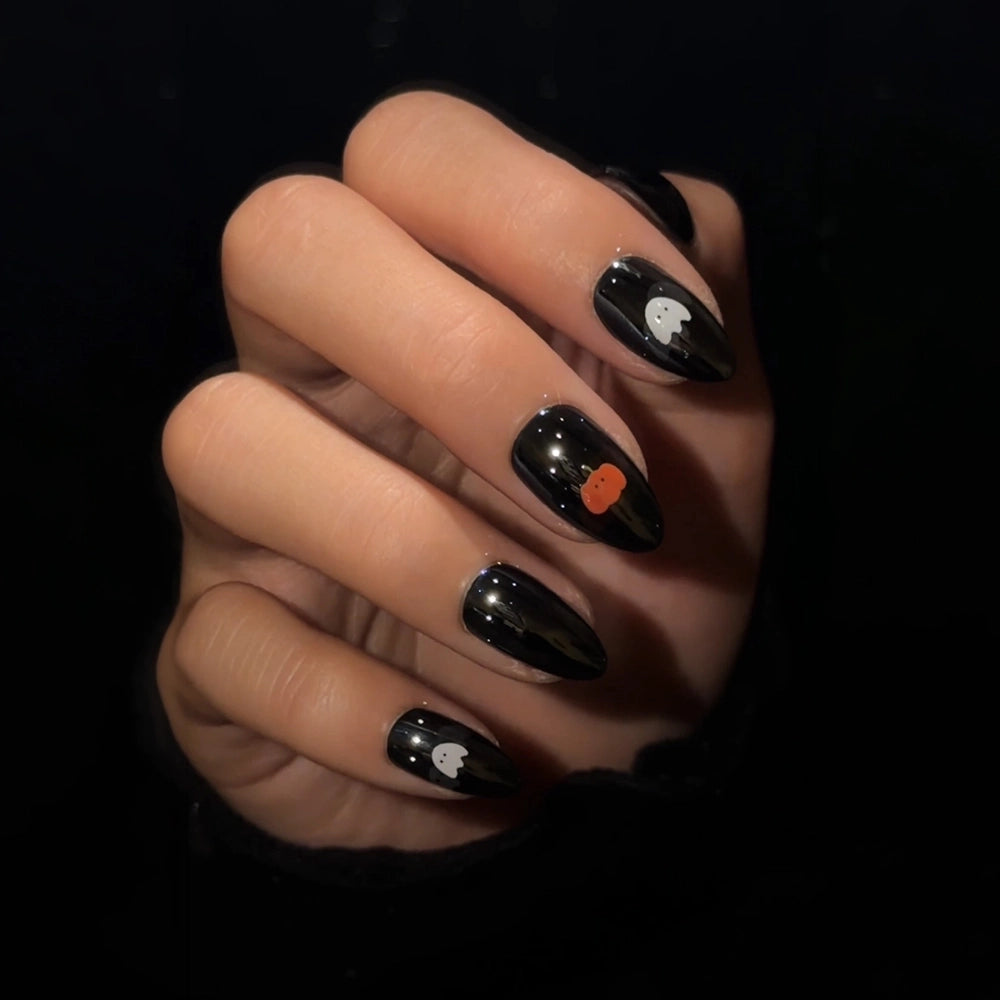 A hand with long, black, glossy nails featuring a glazed finish showcases the “Tricks & Treats” press-on nail set by glamnetic. Two nails display small white ghost designs, while another features a tiny orange pumpkin—all set against a dark background for a striking Halloween-inspired look that’s easy to achieve. - Product image