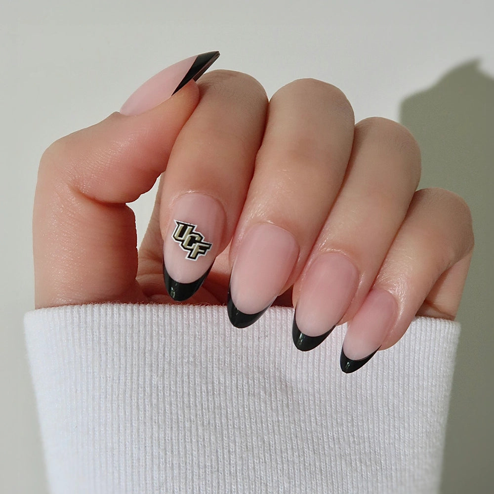 A hand wearing the glamnetic UCF Knights Press-On Nail Set features almond-shaped nails with black French tips, a nude base, and the team's logo on the ring finger. The person is dressed in a white long-sleeve shirt. - Product image