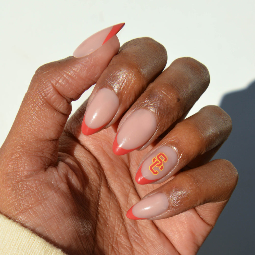 A hand with almond-shaped Collegiate Press-On Nails by glamnetic is shown: the nails are painted nude with red tips, and the ring fingernail features a yellow and red USC logo. The background is plain white. - Product image