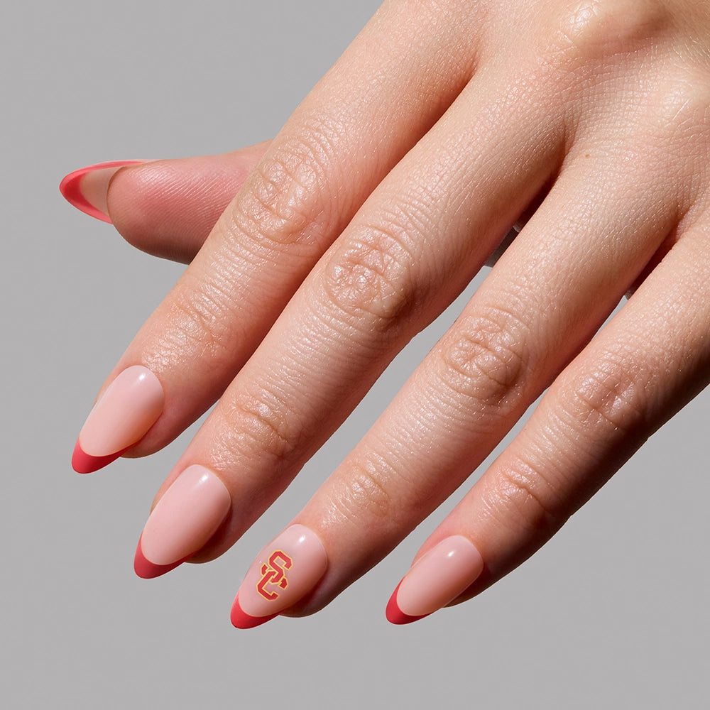 A hand with almond-shaped nude nails featuring red French tips wears the glamnetic USC Trojans Press-On Nail Set. The ring finger displays a yellow and red USC logo against a plain gray background. - Product image
