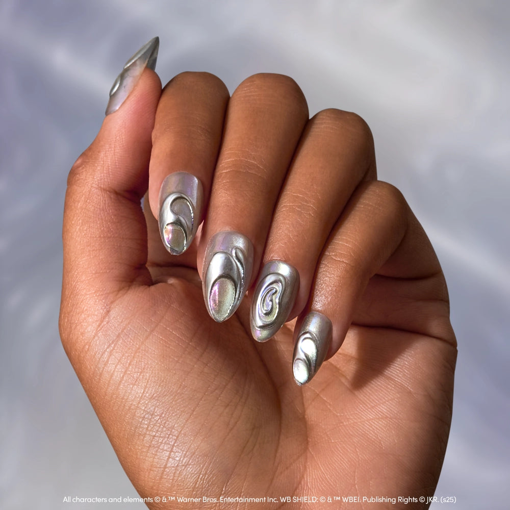 A hand shows long, almond-shaped chrome nails painted in glamnetic's Unicorn Blood, a metallic silver polish. Some feature raised, 3D silver art for a futuristic look that evokes magical creatures against a soft, blurred background. - Product image