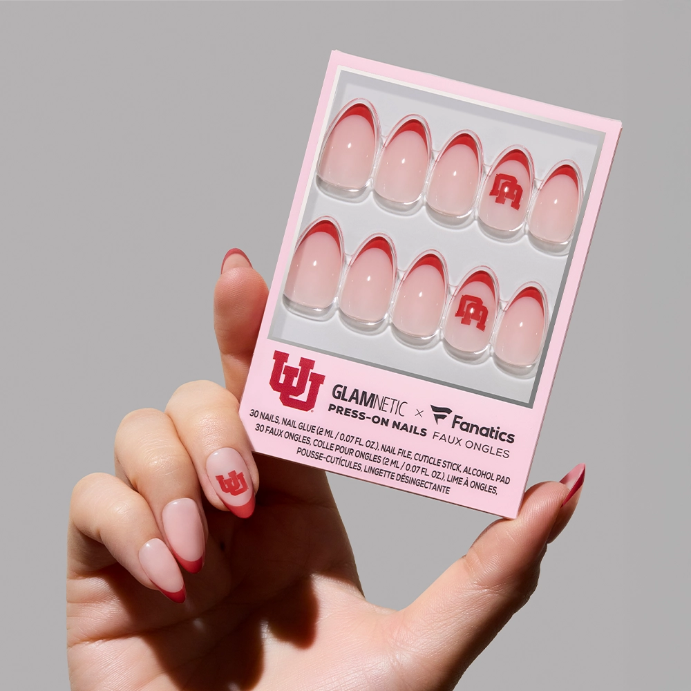 A hand holds the glamnetic Utah Utes Press-On Nail Set, featuring red and pink French tips with the University of Utah logo. The packaging highlights salon-quality nails from glamnetic. - Product image