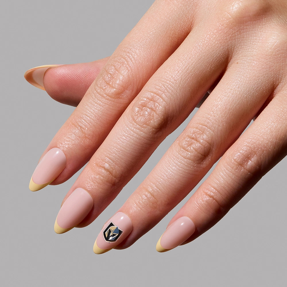 A hand with long, almond-shaped nails sports the glamnetic Vegas Golden Knights Press-On Nail Set in light pink with pale yellow tips and a silver geometric charm on the ring finger, perfect for fandom style against a plain gray background. - Product image