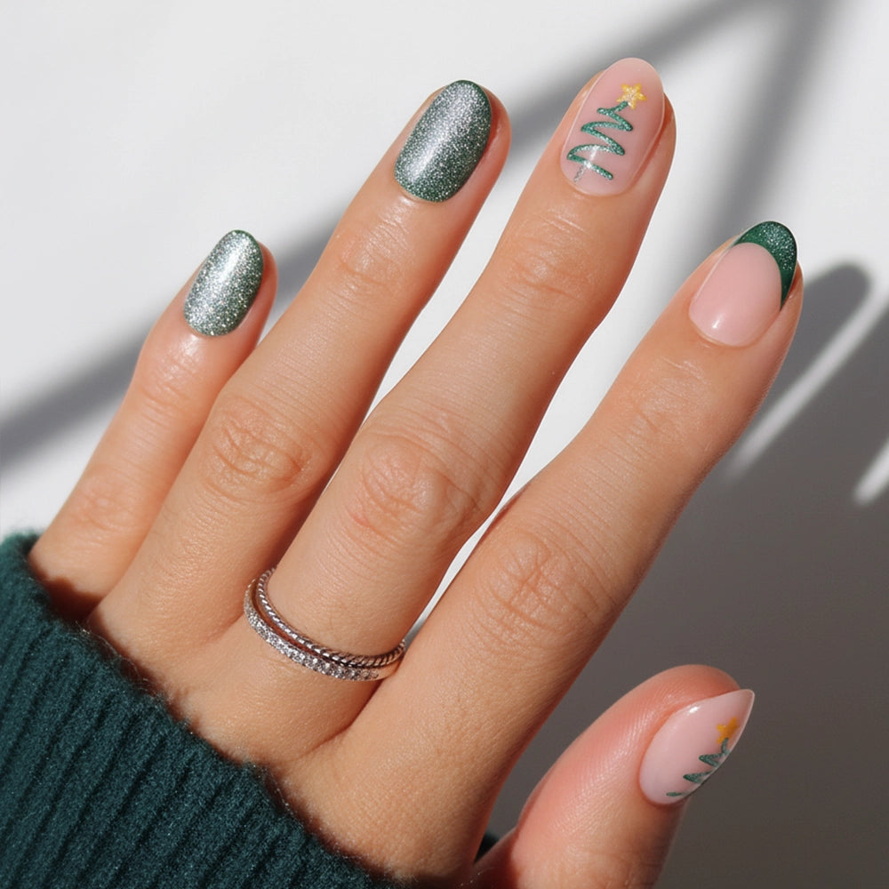 A hand shows off a festive holiday manicure featuring glamnetic’s Velvet Pine: green glitter polish, green French tips, and Christmas tree designs on short nude oval nails. The look is accessorized with two silver rings and a green sweater.
