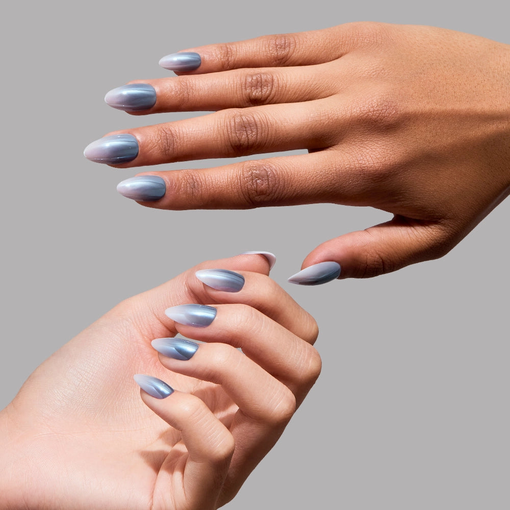 Two hands with long, almond-shaped nails painted in glamnetic’s Vintage Silver, a striking metallic blue-gray shade, are shown against a plain gray background. Both hands are elegantly posed to showcase the nails, highlighting the sophisticated glazed finish of this set. - Product image