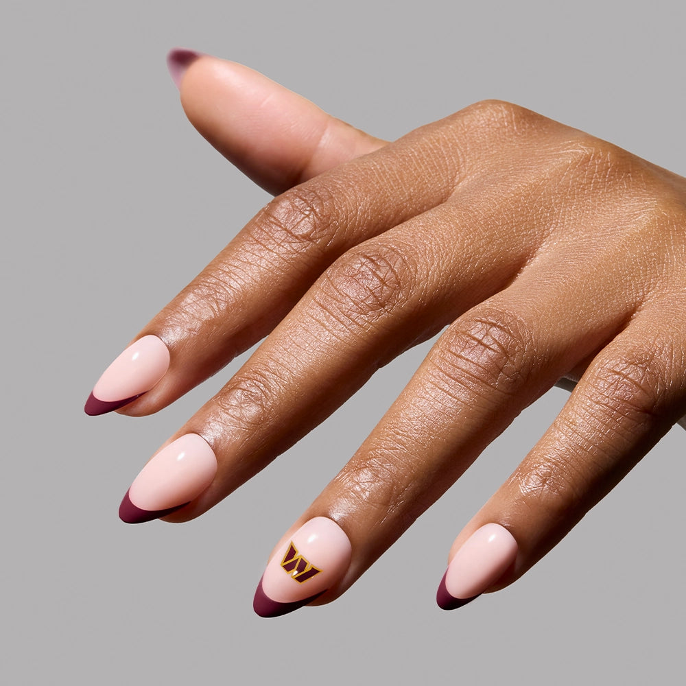 The glamnetic Washington Commanders Press-On Nail Set features almond-shaped nails with a pink base, burgundy tips, and a gold geometric accent on the ring finger, all set against a simple light gray background. - Product image