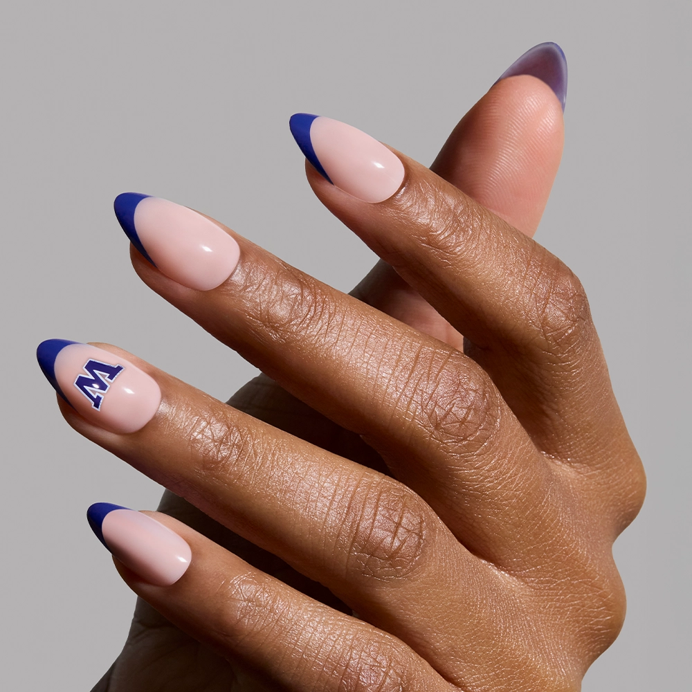 A hand with almond-shaped nails wears the glamnetic Washington Huskies Press-On Nail Set, featuring a nude base, dark blue French tips, and a blue and white "N" decal; shown against a plain light gray background. - Product image