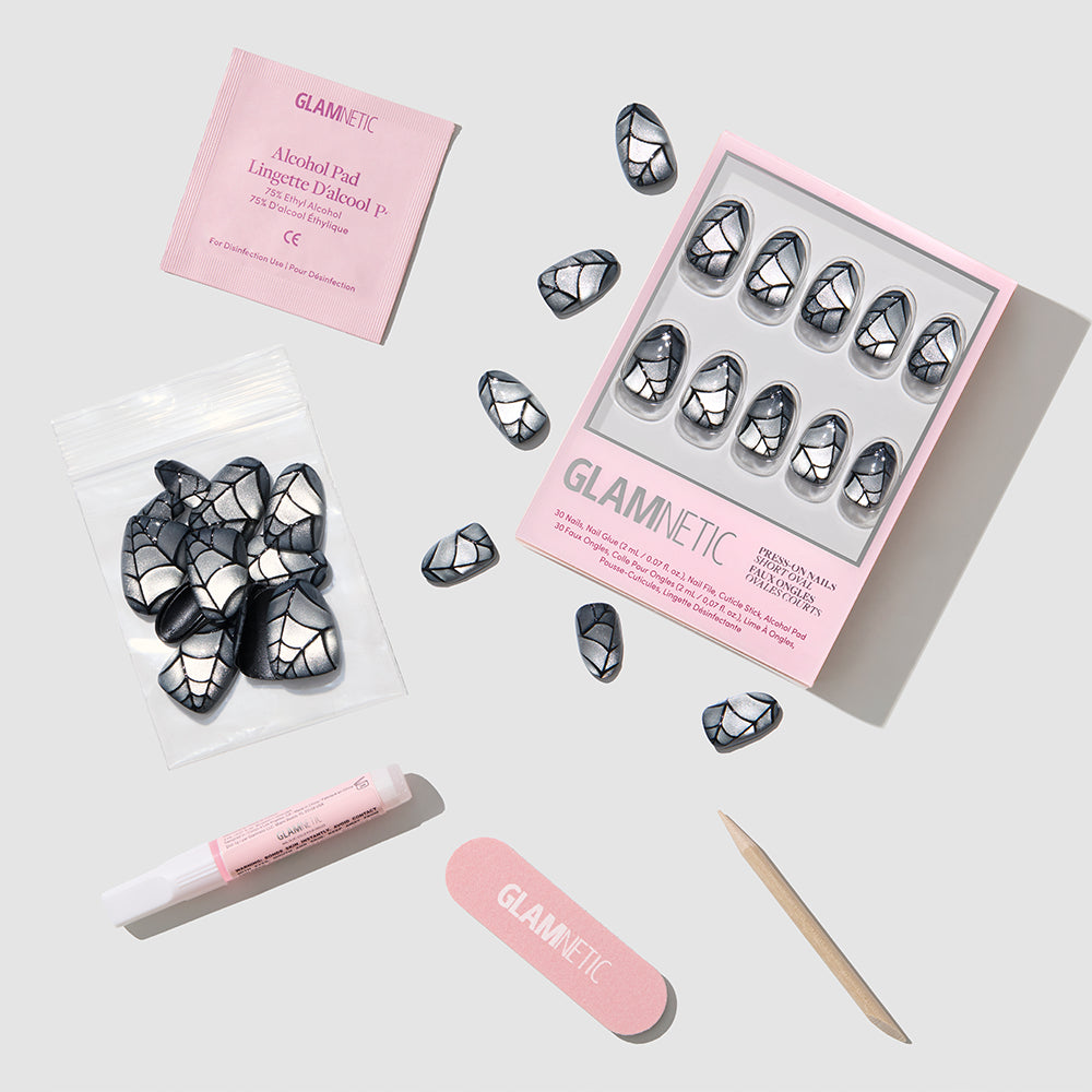 Glamnetic press-on nail kit featuring trendy short oval nails with a black and silver cracked pattern reminiscent of chic black velvet nails. The set includes a box of 30 press-on nails, extra nails in a plastic bag, nail glue, a nail file, a cuticle stick, and an alcohol pad—all displayed on a sleek gray background. - Product image